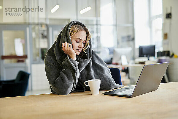 Erschöpfte junge Frau in gemütlicher Loungewear sitzt mit Laptop am Schreibtisch im Büro