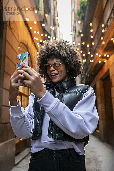Glückliche junge Frau mit Sonnenbrille  die in einer Gasse ein Selfie mit dem Smartphone macht