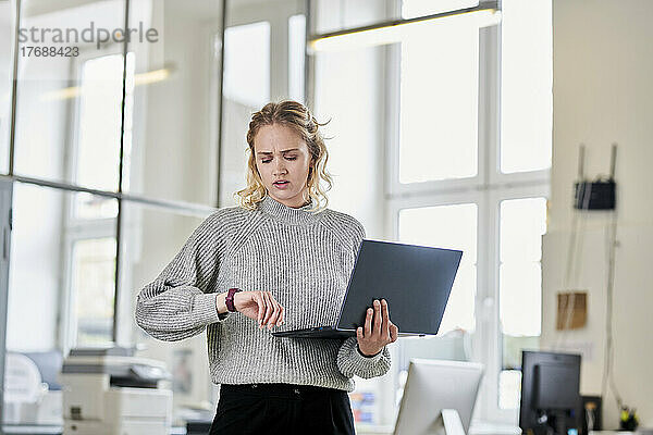 Junge Frau steht mit Laptop im Büro und schaut auf die Uhrzeit