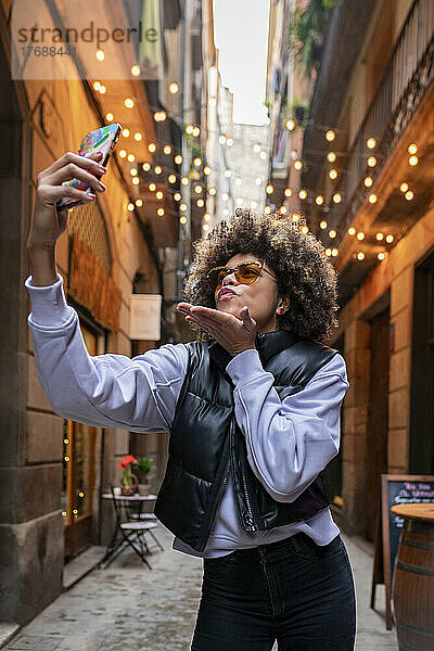 Junge Frau bläst Kuss und macht Selfie mit dem Handy in einer Gasse