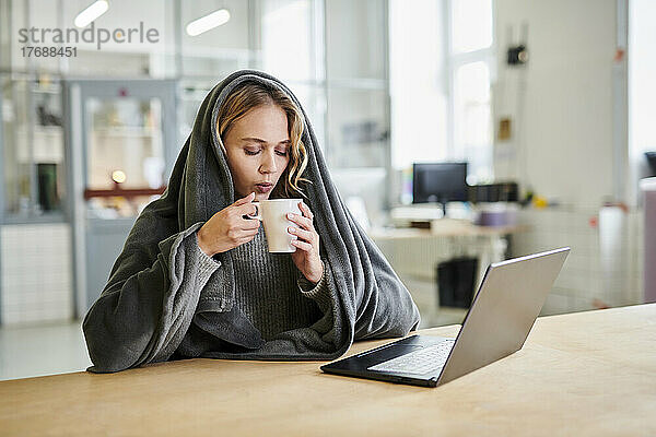 Junge Frau in gemütlicher Loungewear sitzt am Schreibtisch im Büro und trinkt aus einer Tasse