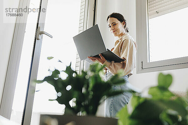 Smiling young woman standing with laptop by door at home