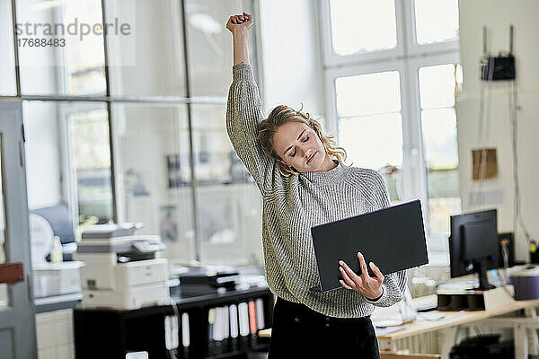 Müde junge Frau steht im Büro und hält Laptop in der Hand