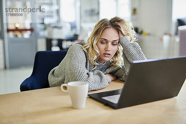 Müde junge Frau sitzt mit Laptop am Schreibtisch im Büro
