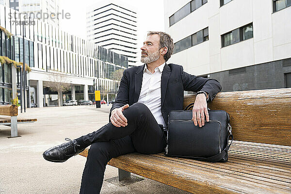 Reifer Geschäftsmann mit Laptoptasche sitzt auf Bank vor Bürogebäude