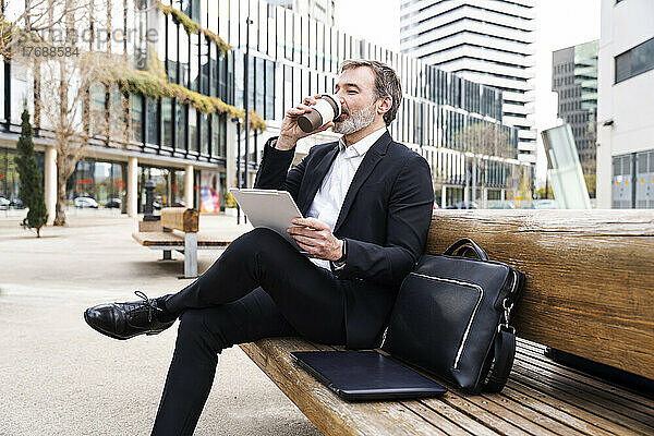 Geschäftsmann trinkt Kaffee und sitzt mit Tablet-PC auf einer Bank vor dem Bürogebäude