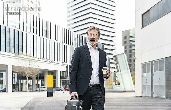 Geschäftsmann mit Laptoptasche und Einwegkaffeetasse steht vor Bürogebäude