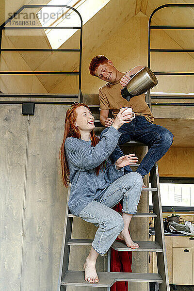 Lächelnder Junge mit Freundin sitzt auf Leiter und gießt Tee in Tasse