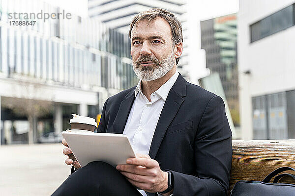 Reifer Geschäftsmann mit Tablet-PC und Einweg-Kaffeetasse sitzt auf der Bank