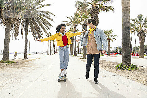 Glücklicher Mann und Frau beim Skateboarden vor Palmen auf dem Fußweg