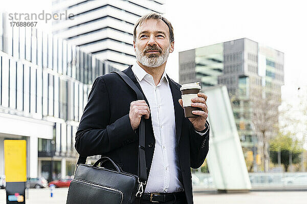 Lächelnder reifer Geschäftsmann mit Laptoptasche und Einweg-Kaffeetasse