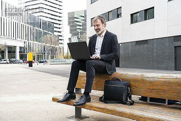 Lächelnder Geschäftsmann sitzt mit Laptop auf Bank vor Bürogebäude