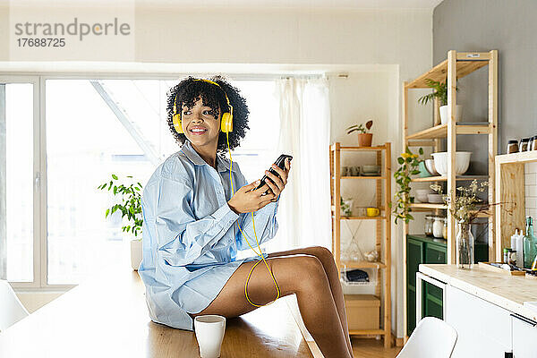 Lächelnde junge Frau mit Kopfhörern sitzt zu Hause auf der Kücheninsel