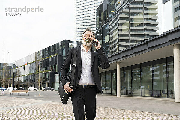 Lächelnder reifer Geschäftsmann mit Laptoptasche  der auf dem Smartphone spricht und vor dem Bürogebäude läuft