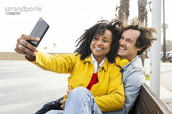 Lächelnde Frau und Mann  die auf einer Holzbank sitzen und ein Selfie mit dem Mobiltelefon machen