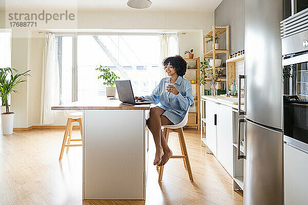 Glückliche Frau sitzt mit Laptop auf einem Stuhl auf der Kücheninsel