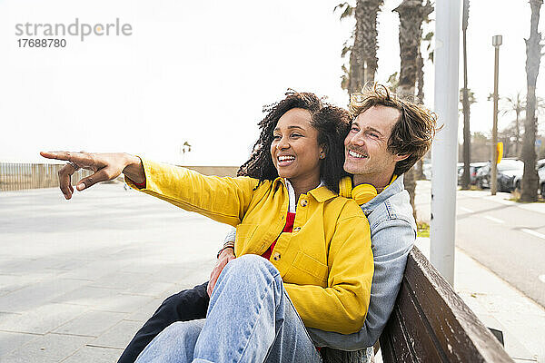 Glückliche Frau sitzt auf Holzbank und Mann zeigt auf Fußweg