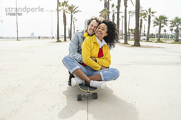 Glücklicher Mann umarmt Frau  die auf Skateboard vor Bäumen sitzt