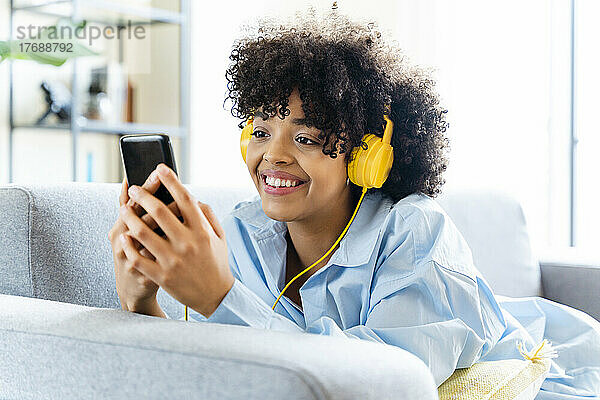 Glückliche Frau mit Kopfhörern und Smartphone  die zu Hause auf dem Sofa liegt