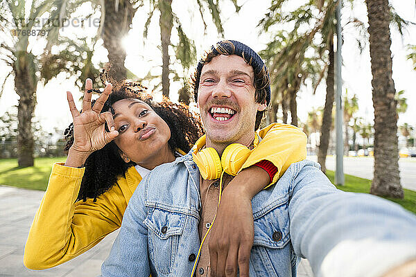 Fröhlicher Mann mit Kopfhörern neben Frau  die im Park ein „OK“-Schild zeigt