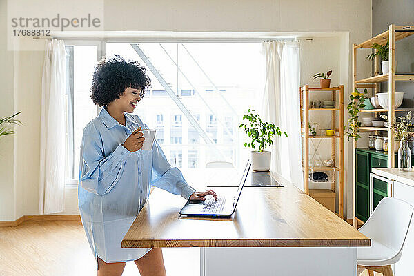Glückliche Frau mit Kaffeetasse und Laptop auf der Kücheninsel