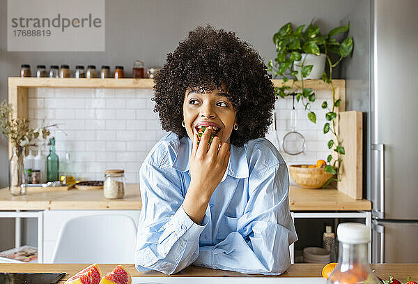 Schöne Frau isst Erdbeeren und lehnt sich zu Hause an die Kücheninsel
