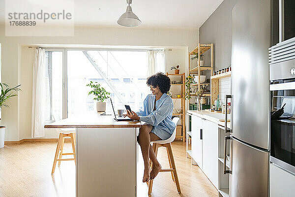 Lächelnde Frau mit Smartphone und Laptop sitzt auf einem Stuhl auf der Kücheninsel