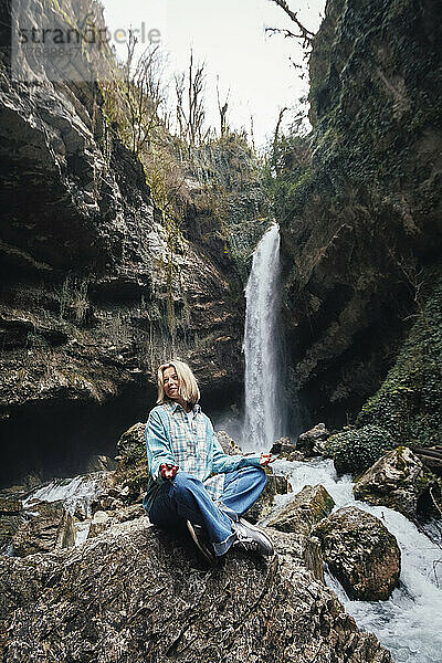 Reife Frau sitzt auf einem Felsen vor dem Wasserfall