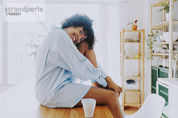 Glückliche Frau mit Kaffeetasse sitzt zu Hause auf der Kücheninsel