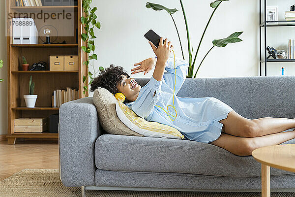 Lächelnde Frau winkt bei einem Videoanruf über ihr Smartphone auf dem heimischen Sofa