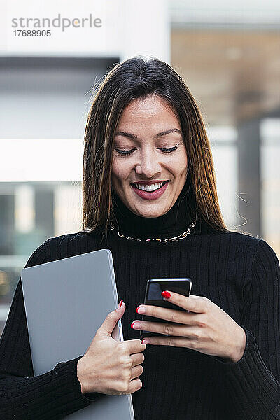 Lächelnde junge Geschäftsfrau mit braunen Haaren  die ihr Smartphone benutzt