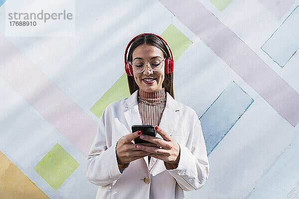 Lächelnde junge Frau mit kabellosen Kopfhörern und Mobiltelefon vor der Wand