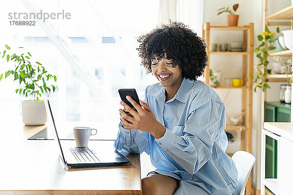 Glückliche junge Frau sitzt auf der Kücheninsel und benutzt ihr Smartphone