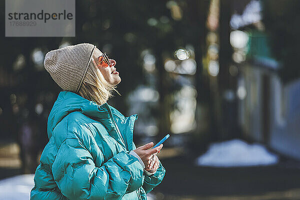 Lächelnde Frau mit Sonnenbrille und Smartphone an einem sonnigen Tag