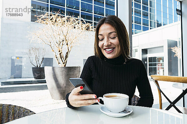 Lächelnde Geschäftsfrau mit Kaffeetasse und Smartphone im Café