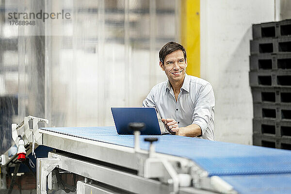 Smiling businessman with laptop on conveyor belt in warehouse