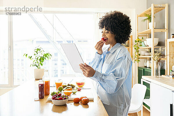 Junge Frau benutzt Tablet-PC und isst Erdbeeren in der heimischen Küche
