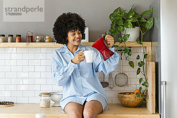 Glückliche Frau  die zu Hause auf der Küchentheke sitzt und Kaffee in eine Tasse gießt