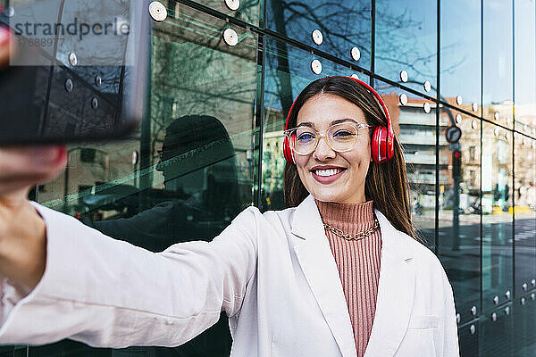 Lächelnde Frau mit kabellosen Kopfhörern  die ein Selfie mit dem Smartphone macht