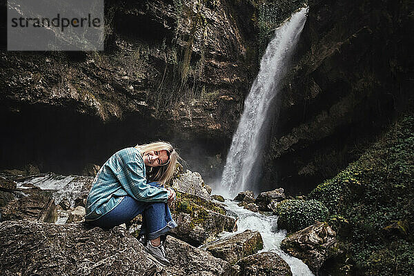 Glückliche reife Frau sitzt auf einem Felsen vor dem Wasserfall