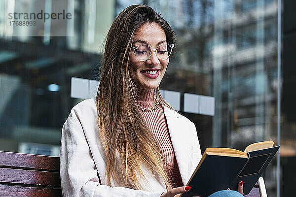 Lächelnde Frau mit Brille sitzt vor einer Glaswand und liest ein Buch