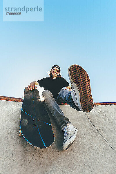 Glücklicher Mann mit Skateboard sitzt im Skateboardpark