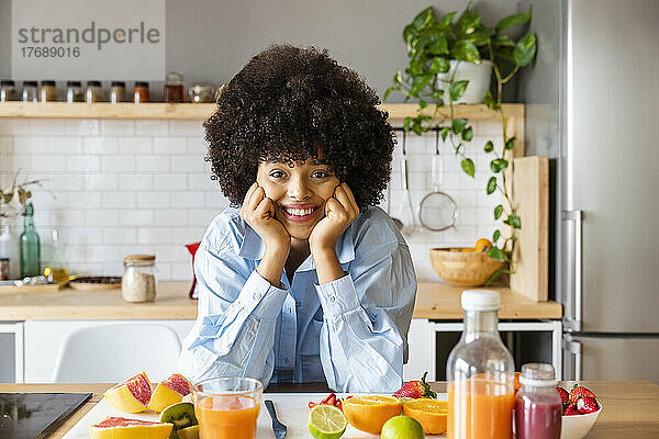 Lächelnde schöne Frau mit Afro-Frisur lehnt an Früchten auf der Kücheninsel zu Hause