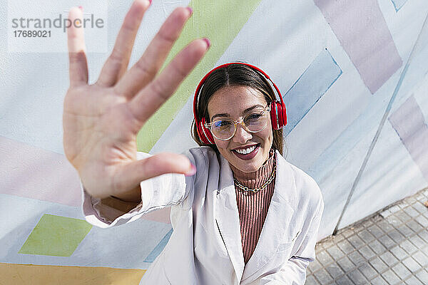 Lächelnde Frau mit Kopfhörern gestikuliert an einem sonnigen Tag mit der Hand vor der Wand