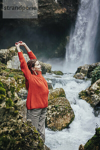 Glückliche junge Frau mit erhobenen Armen steht vor dem Wasserfall