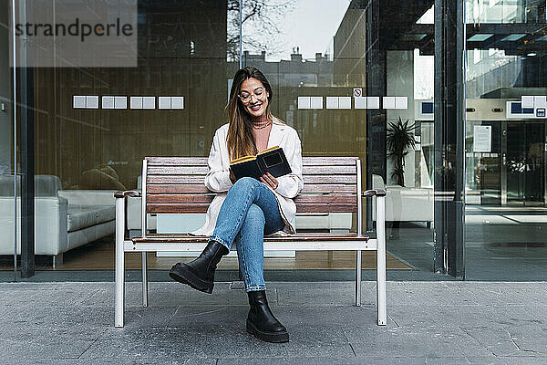 Lächelnde Frau liest ein Buch und sitzt auf einer Bank vor einer Glaswand