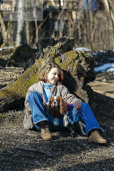 Lächelnde junge Frau mit Hund sitzt an einem sonnigen Tag im Wald
