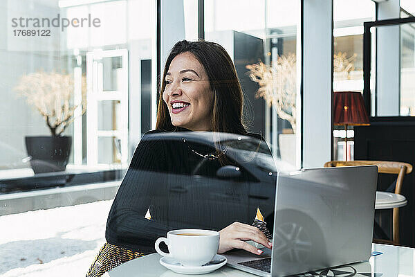 Glückliche Geschäftsfrau mit Laptop  die durch die Glasscheibe im Café gesehen wird