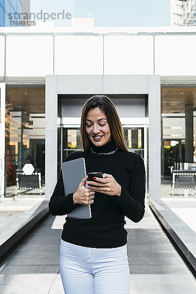 Lächelnde junge Geschäftsfrau mit Laptop und Smartphone vor dem Café