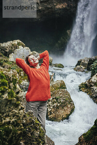 Happy young woman with hands behind head standing in front of waterfall